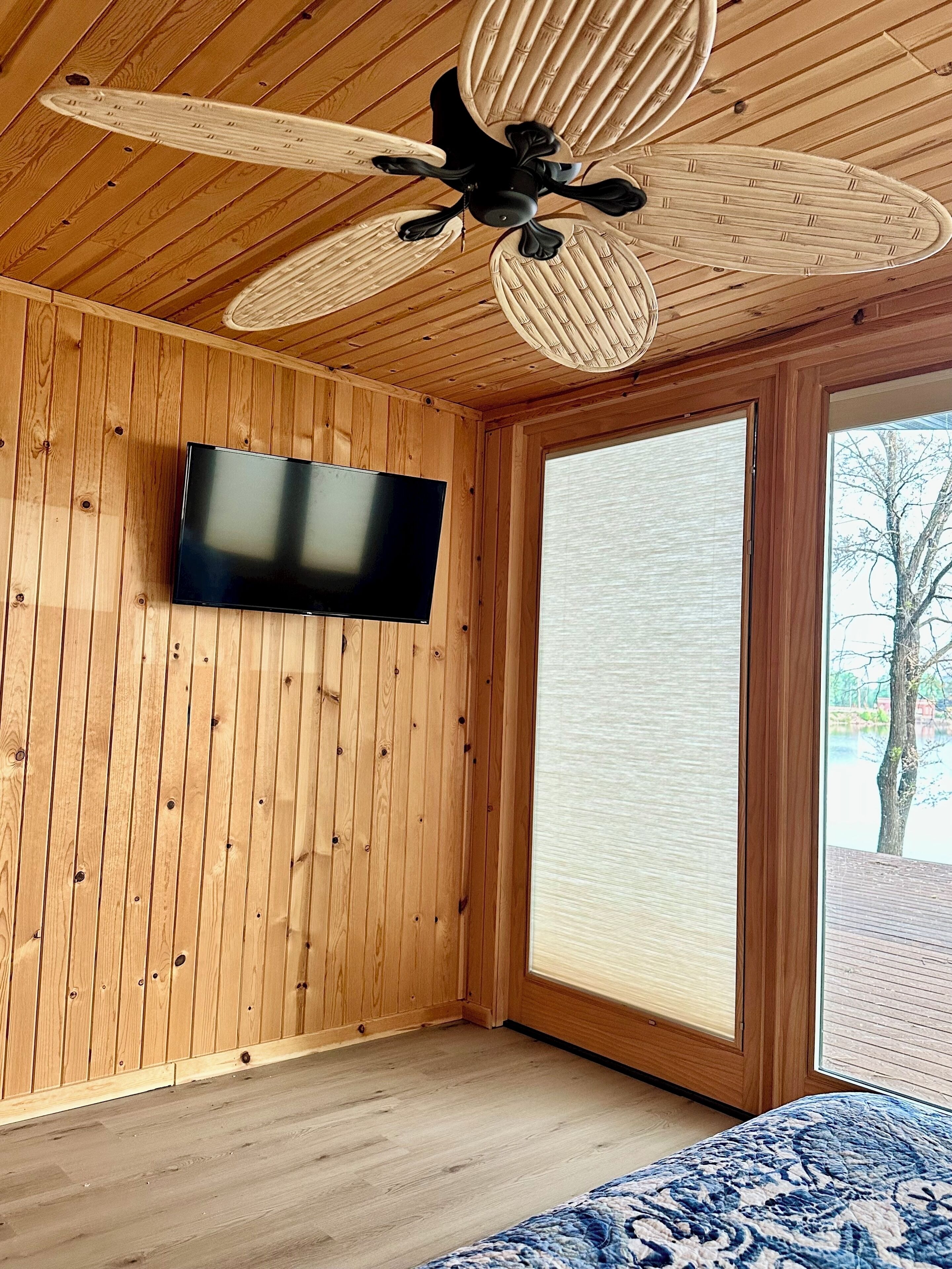 Ein Zimmer mit Holzwänden, einem Deckenventilator, einem wandmontierten Fernseher und einem großen Fenster mit Blick auf Bäume und ein Gewässer.