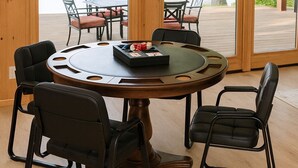 Een ronde houten tafel met zes stoelen, een dienblad met speelkaarten, en uitzicht op een meer door grote ramen.
