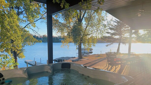 Een hottub met uitzicht op een meer en een steiger, omringd door bomen en een houten terras met stoelen.