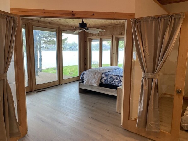 Un dormitorio con cama, ventilador de techo y amplios ventanales con vistas a un lago.