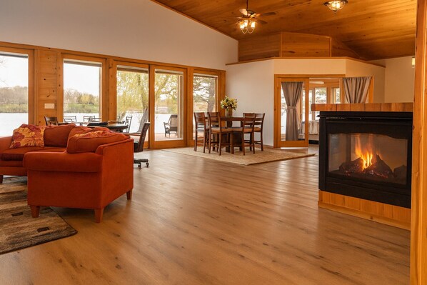 A spacious living room with a fireplace, wooden floors, and large windows offering a view of the outdoors.
