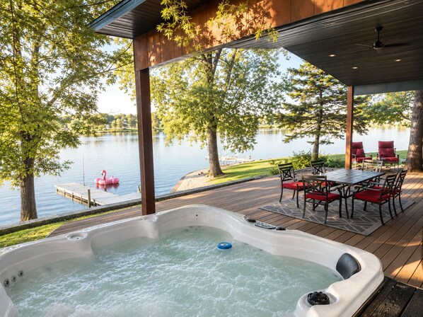 Un jacuzzi en una terraza de madera con vista a un lago y un muelle con un flotador con forma de flamenco.