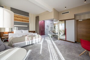 Habitación de hotel moderna con una cama grande, zona de ducha separada, baño con paredes de vidrio y una silla roja.