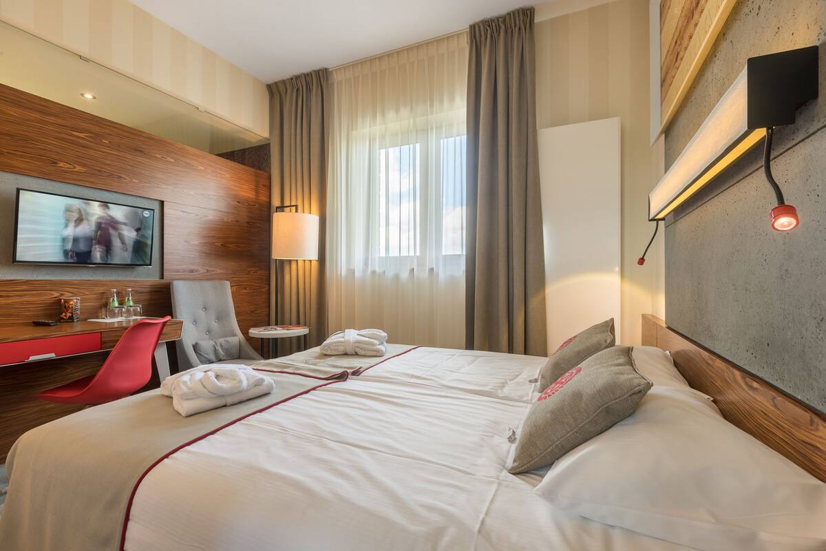 Habitación de hotel con una cama grande, un televisor montado en la pared, un escritorio con una silla y una ventana con cortinas.