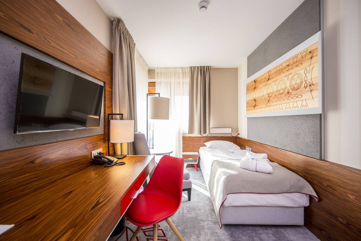 Habitación de hotel con un escritorio de madera, una silla roja, una cama con ropa blanca, un televisor montado en la pared y un cuadro abstracto grande en la pared.