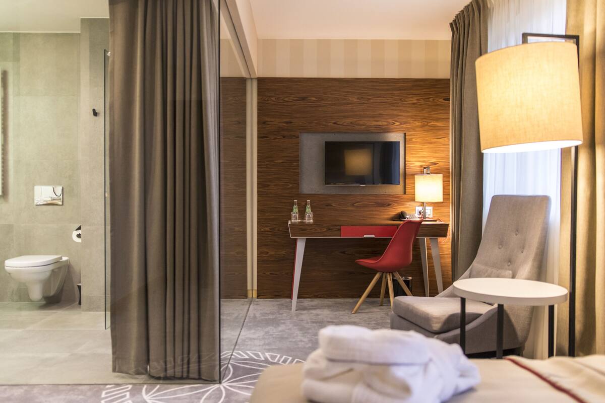 Habitación de hotel con pared de acento de madera, un televisor de pantalla plana sobre un escritorio, una silla roja, una mesa redonda y un baño con inodoro y ducha.