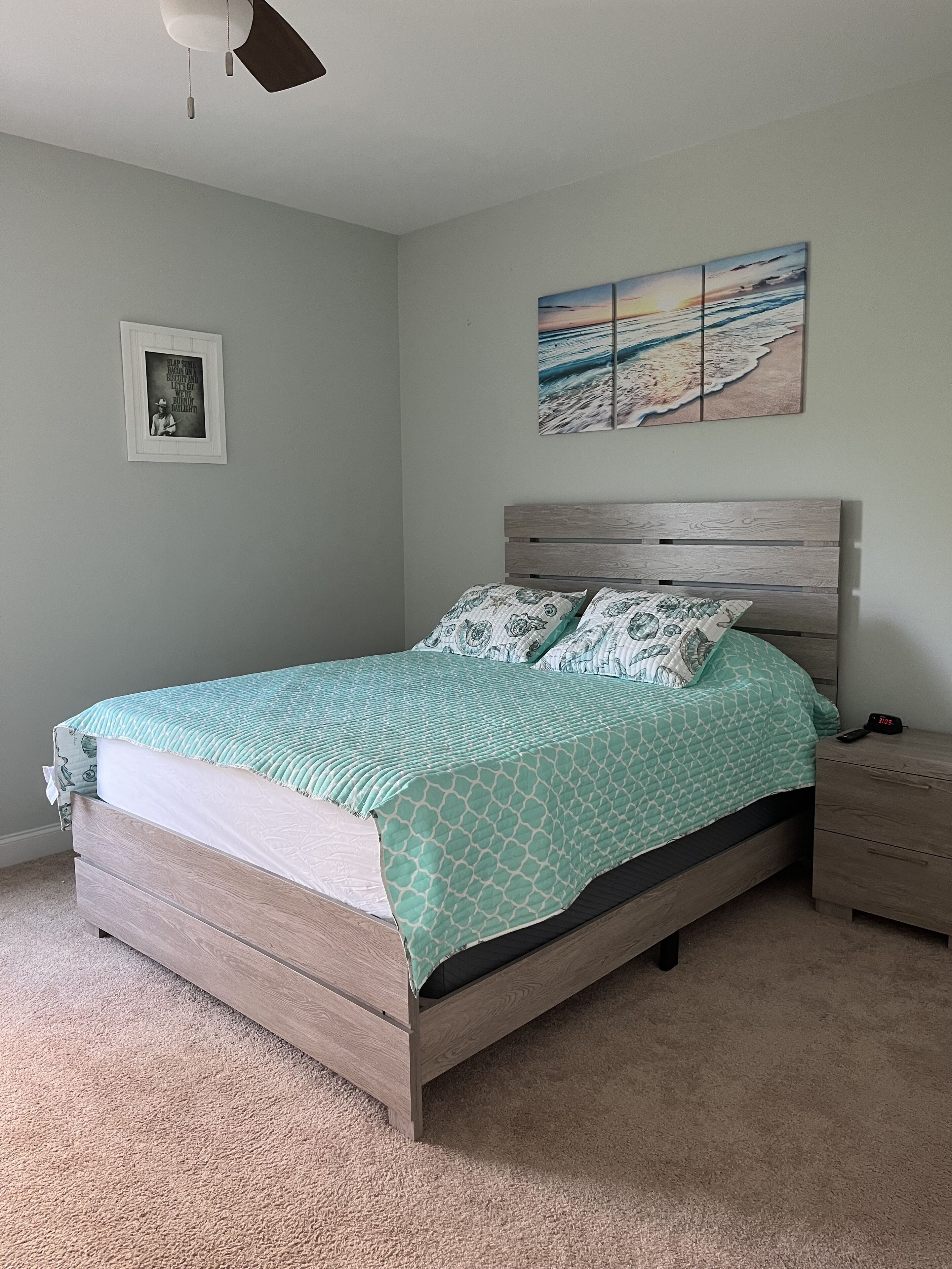 A bedroom with a bed, a nightstand, a wall-mounted painting, and a ceiling fan.