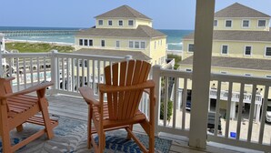 Una terraza de madera con dos sillas Adirondack frente a una playa y vista al océano, con casas al fondo.