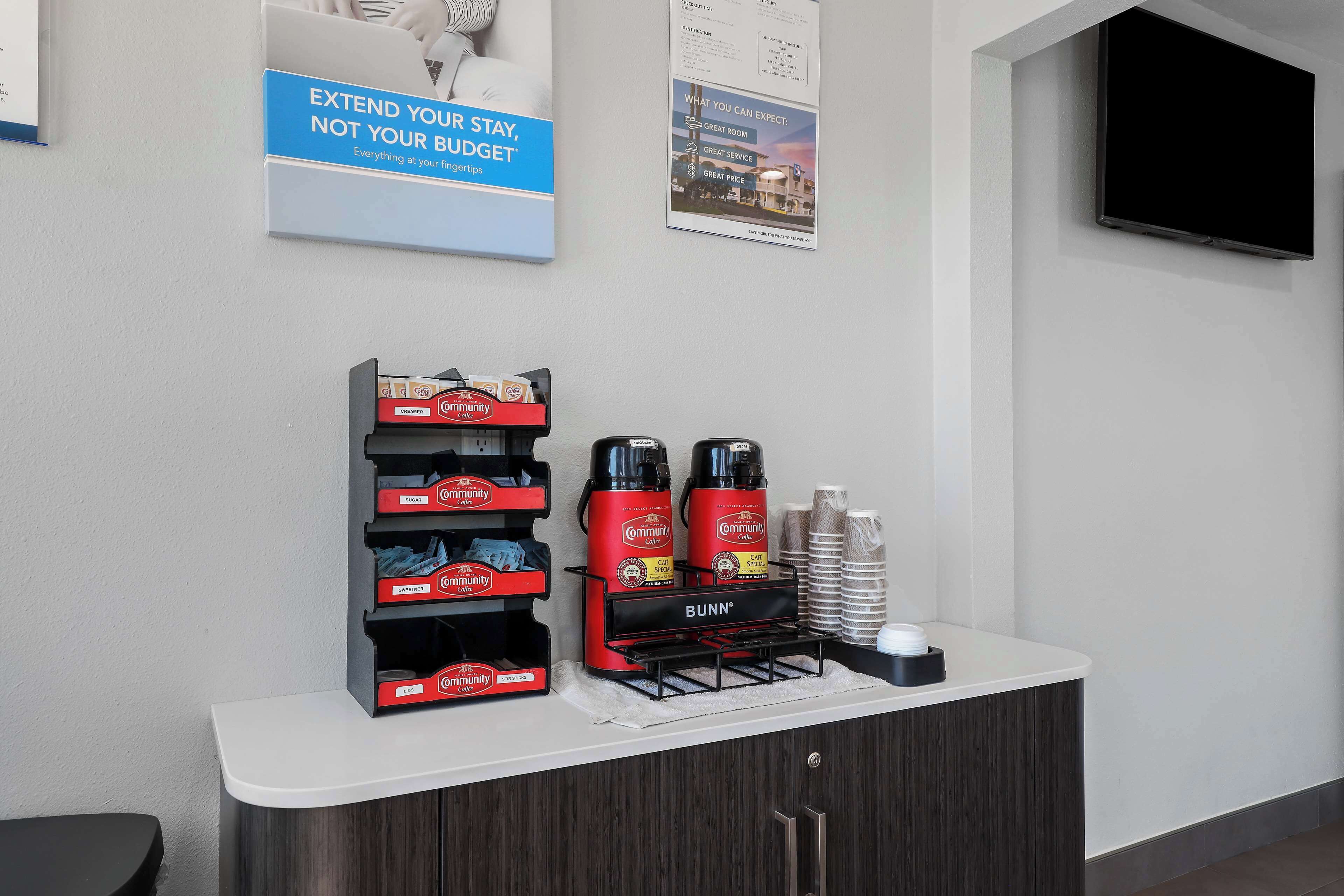 A hotel reception area with a counter displaying coffee makers, cups, and a Bunn coffee machine. There are posters on the wall advertising the hotel's services.
