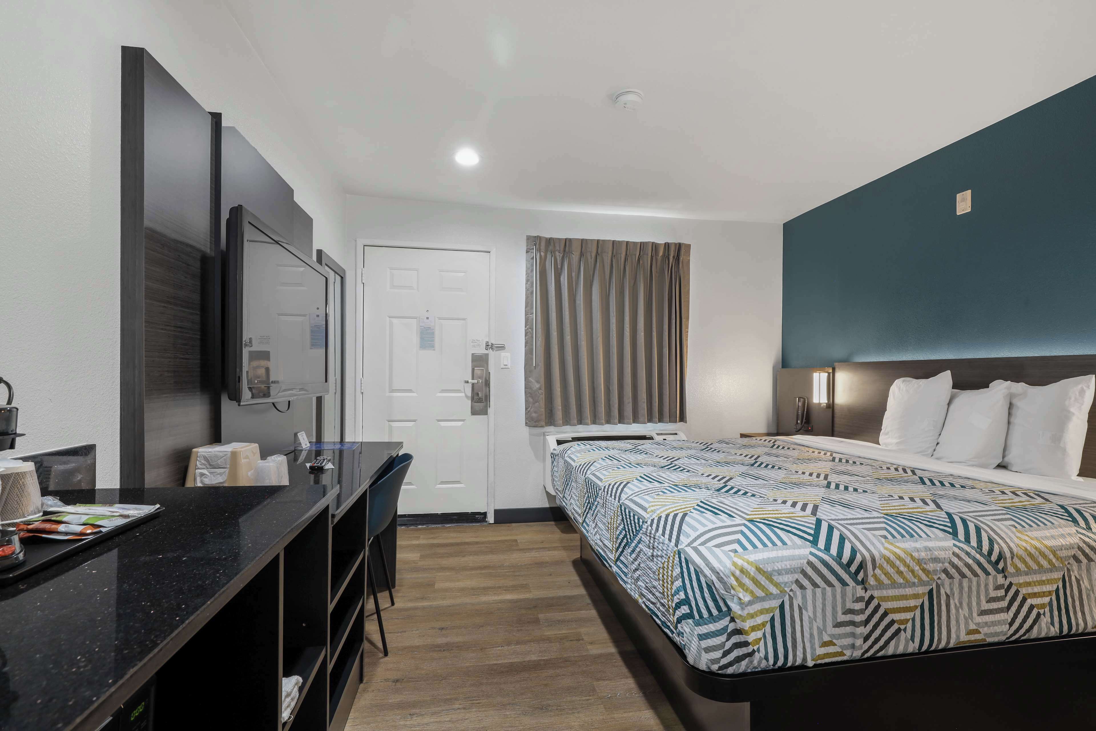 A hotel room with a bed, a desk, a chair, a mirror, a door, and a curtain.