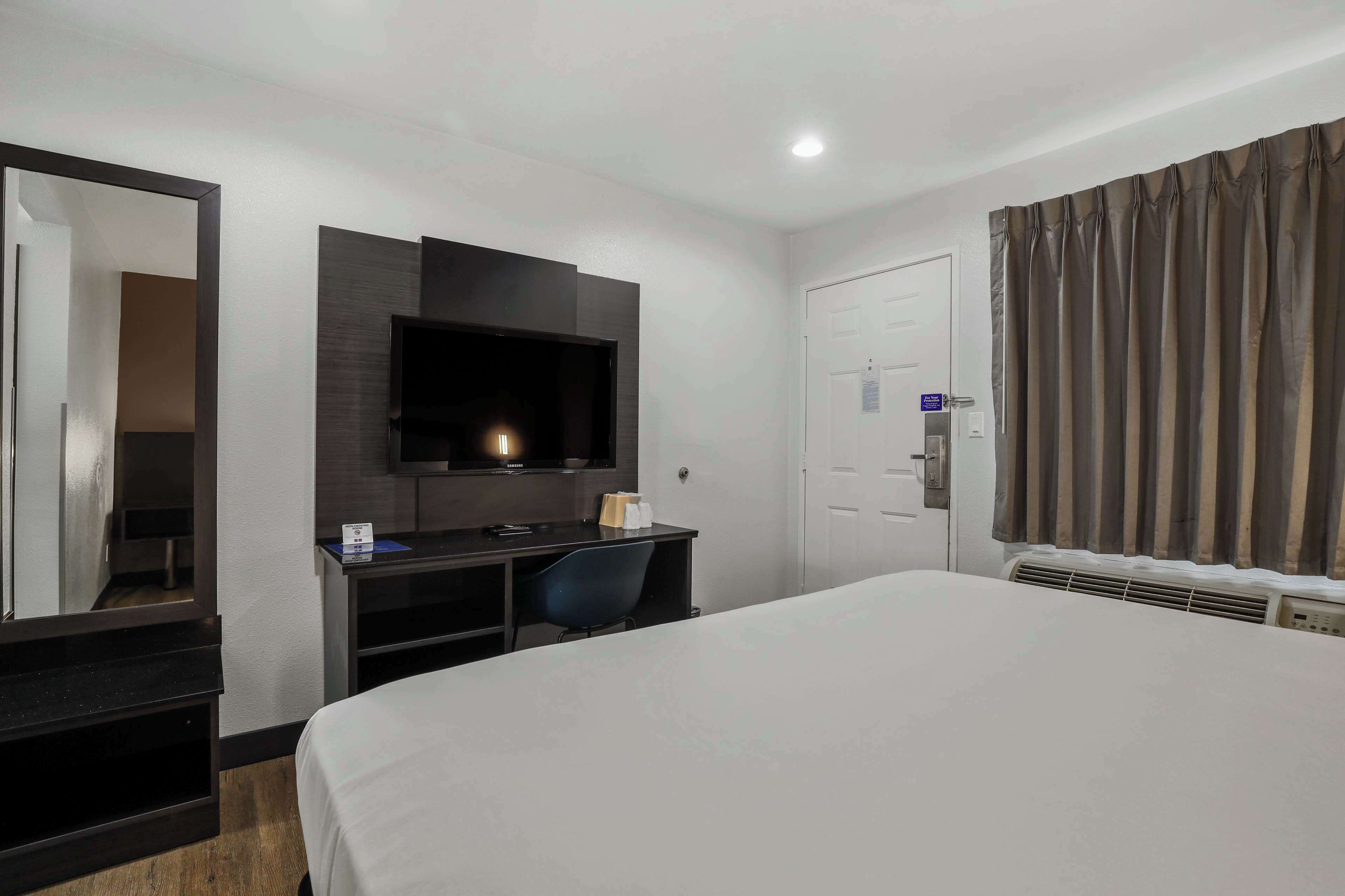 Habitación de hotel con una cama grande, un televisor montado en la pared, un escritorio con una silla y una puerta con un letrero.
