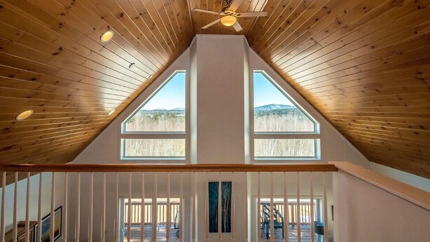 A high-ceilinged room with wooden paneling, a ceiling fan, and two large windows offering a view of the outdoors. - Tranquil mountain view home near downtown North Conway
Mountain Views. Cable TV. (Madison)