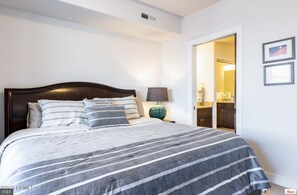 A neatly made bed with a striped bedspread and multiple pillows, a bedside lamp, and a view into a bathroom with a sink and mirror.