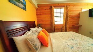 Un dormitorio con cabecera de madera, cama con almohadas blancas y anaranjadas, colcha con estampado floral y ventana con cortinas.