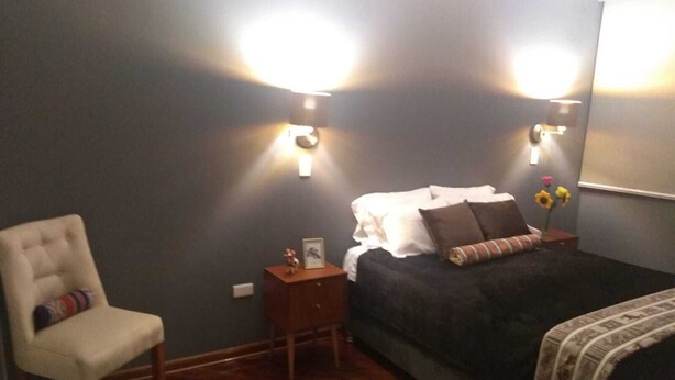 Un dormitorio con una cama, una silla, una mesita de noche y dos lámparas de pared.