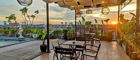 An outdoor patio with wooden flooring, tables, chairs, potted plants, and hanging light fixtures.