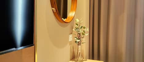 Habitación de hotel con televisión de pantalla plana en la pared, espejo con marco de madera, una mesita con un jarrón de flores y una silla blanca.