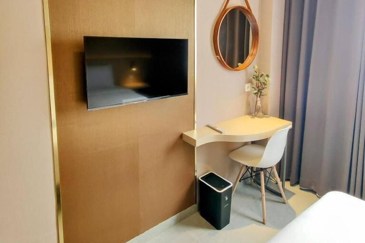 A modern hotel room with a flat-screen TV mounted on the wall, a small desk with a chair, a round mirror, a trash bin, and a bed.