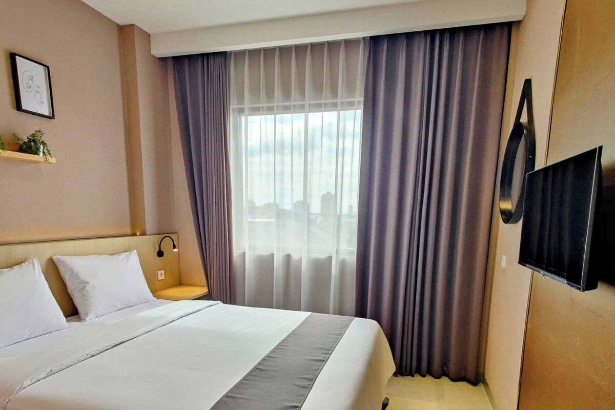 A hotel room with a large bed, a bedside table with a lamp, a TV mounted on the wall, and a window with curtains.