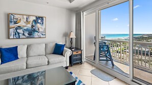 A living room with a sofa, a coffee table, a lamp, and a painting. There is a balcony with a view of the beach.