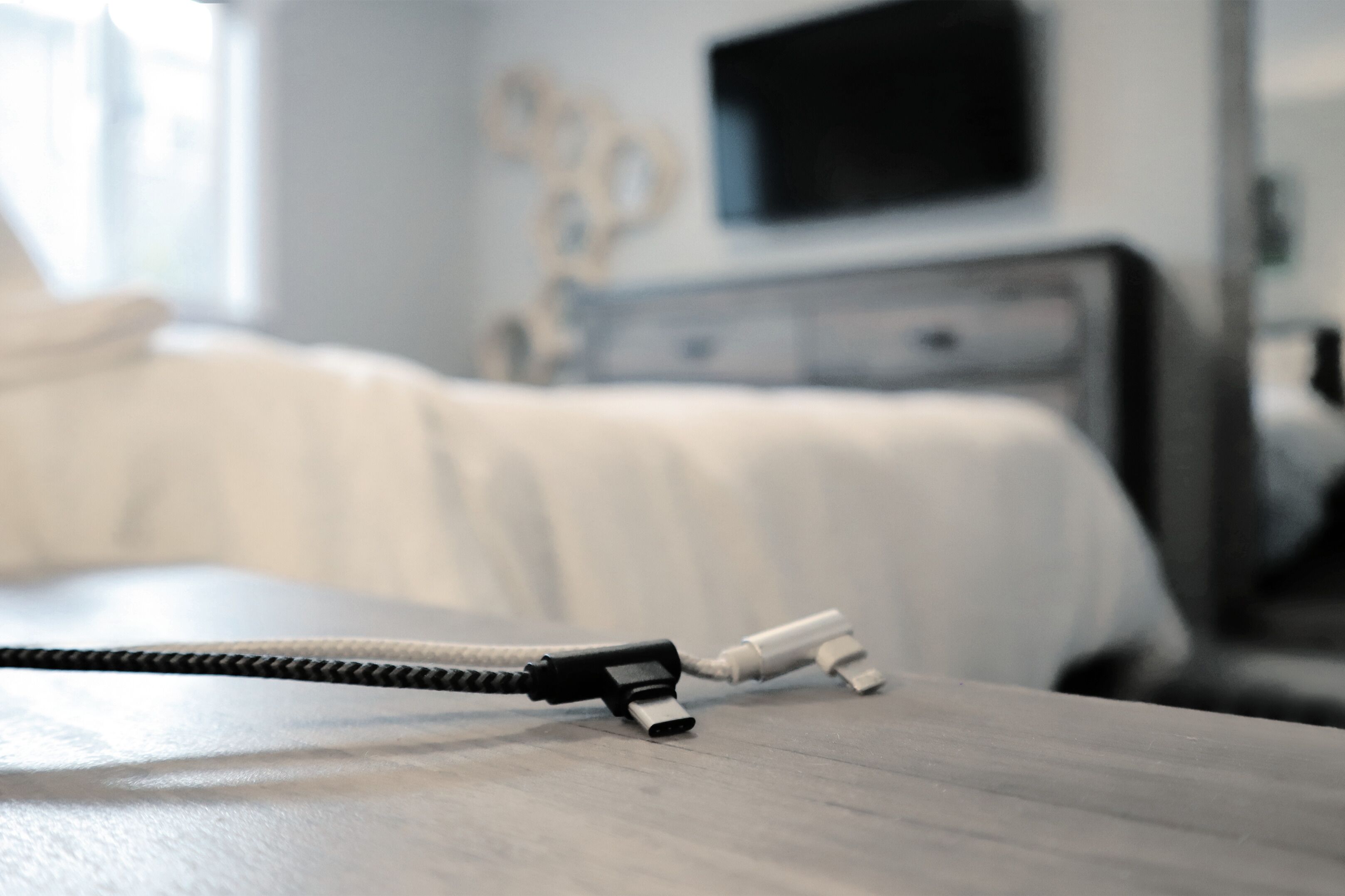 Ein Bett mit weißer Bettwäsche, ein Nachttisch mit Kabel und Netzteil sowie eine Kommode mit einem Flachbildfernseher in einem Hotelzimmer.
