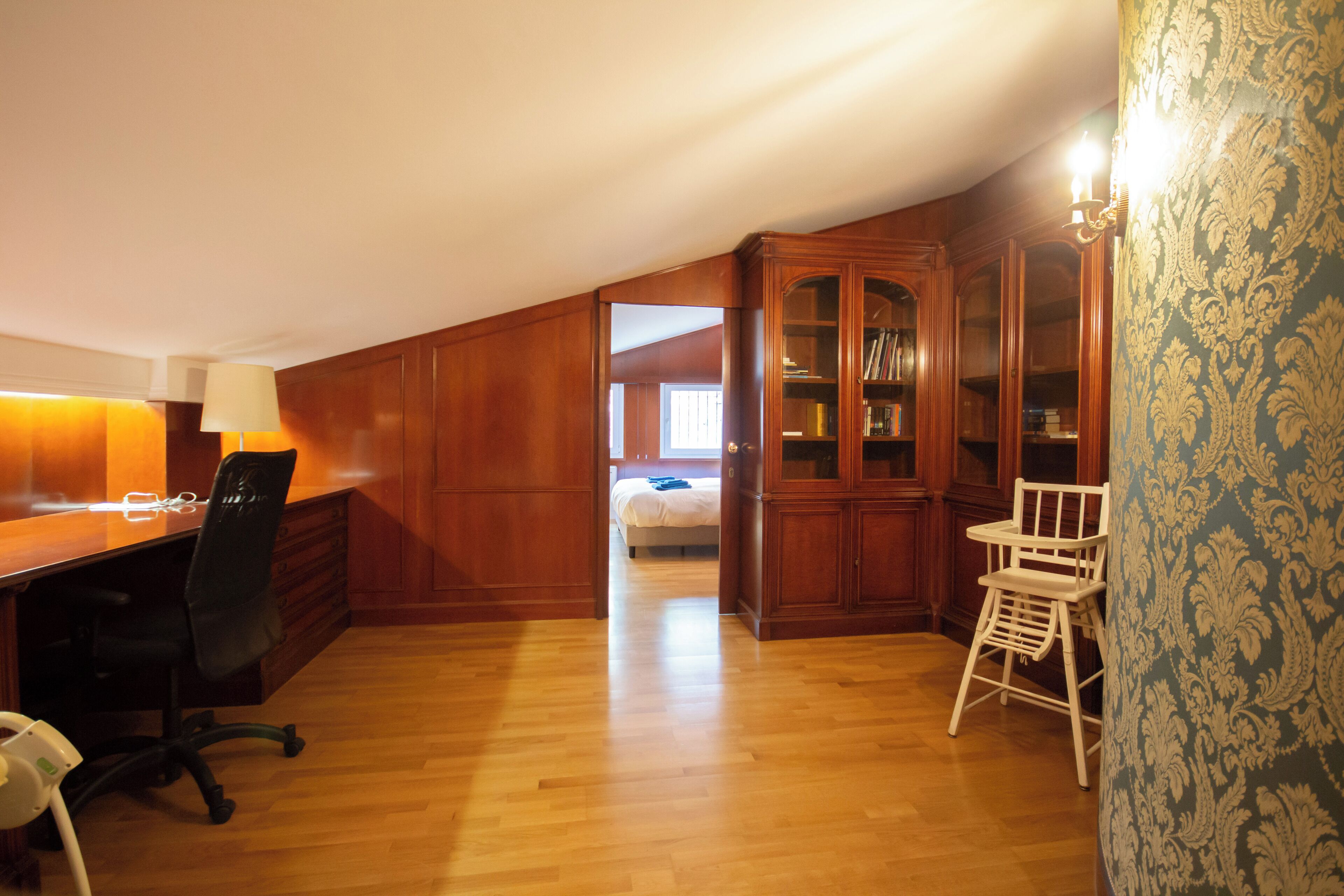 A room with a wooden desk, office chair, and a dining area with a high chair. There is a bookshelf and a bed in the background.