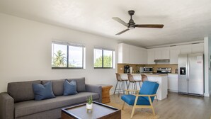 Una sala de estar moderna con un sofá gris, cojines azules, una mesa de centro de madera y una silla azul. Se ve una cocina al fondo, con armarios blancos, un refrigerador de acero inoxidable y una zona de bar.