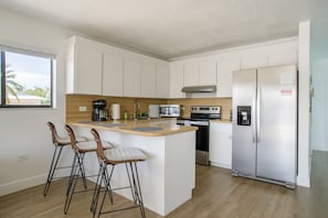 A modern kitchen with white cabinets, a wooden countertop, a stainless steel refrigerator, and a built-in oven. There are three bar stools with cushioned seats.
