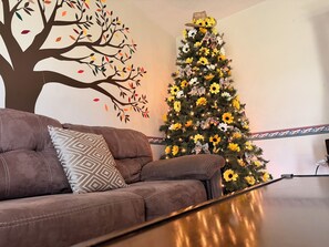 A living room with a brown sofa, a patterned pillow, a wall mural of a tree, and a Christmas tree with yellow flowers and lights. - Cozy Mountain Retreat:The Ultimate WV Getaway w/ large hot tub on spacious porch (Midway)