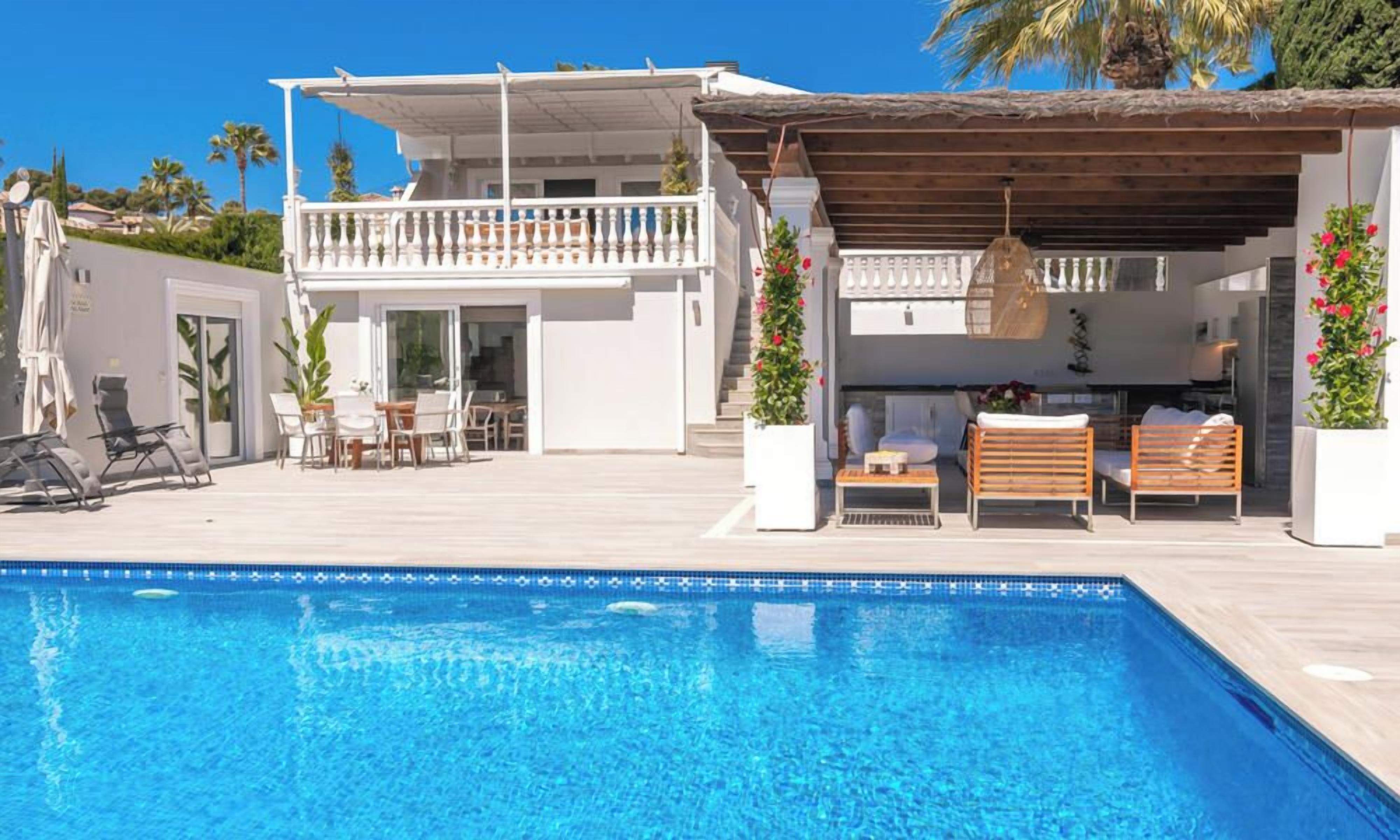 A poolside area with a clear blue pool, a covered seating area with wooden furniture, and a white building with balconies.