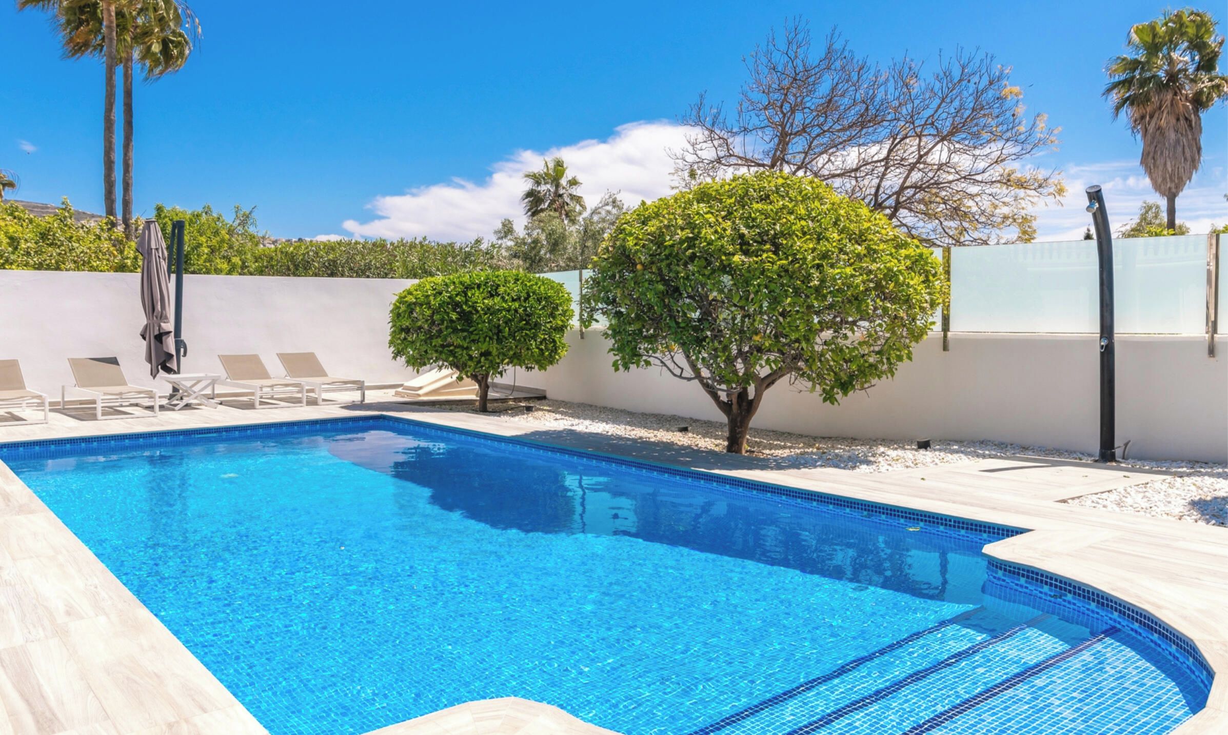A rectangular swimming pool with clear blue water, surrounded by a white wall and a tiled deck.