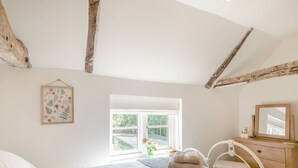 A bedroom with a bed, a window, a bedside table with a mirror, and a framed botanical print on the wall.