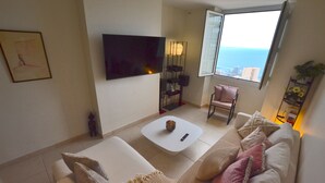 Un salon moderne avec un canapé blanc, une table basse, un téléviseur fixé au mur, un fauteuil et une grande fenêtre donnant sur l’océan.