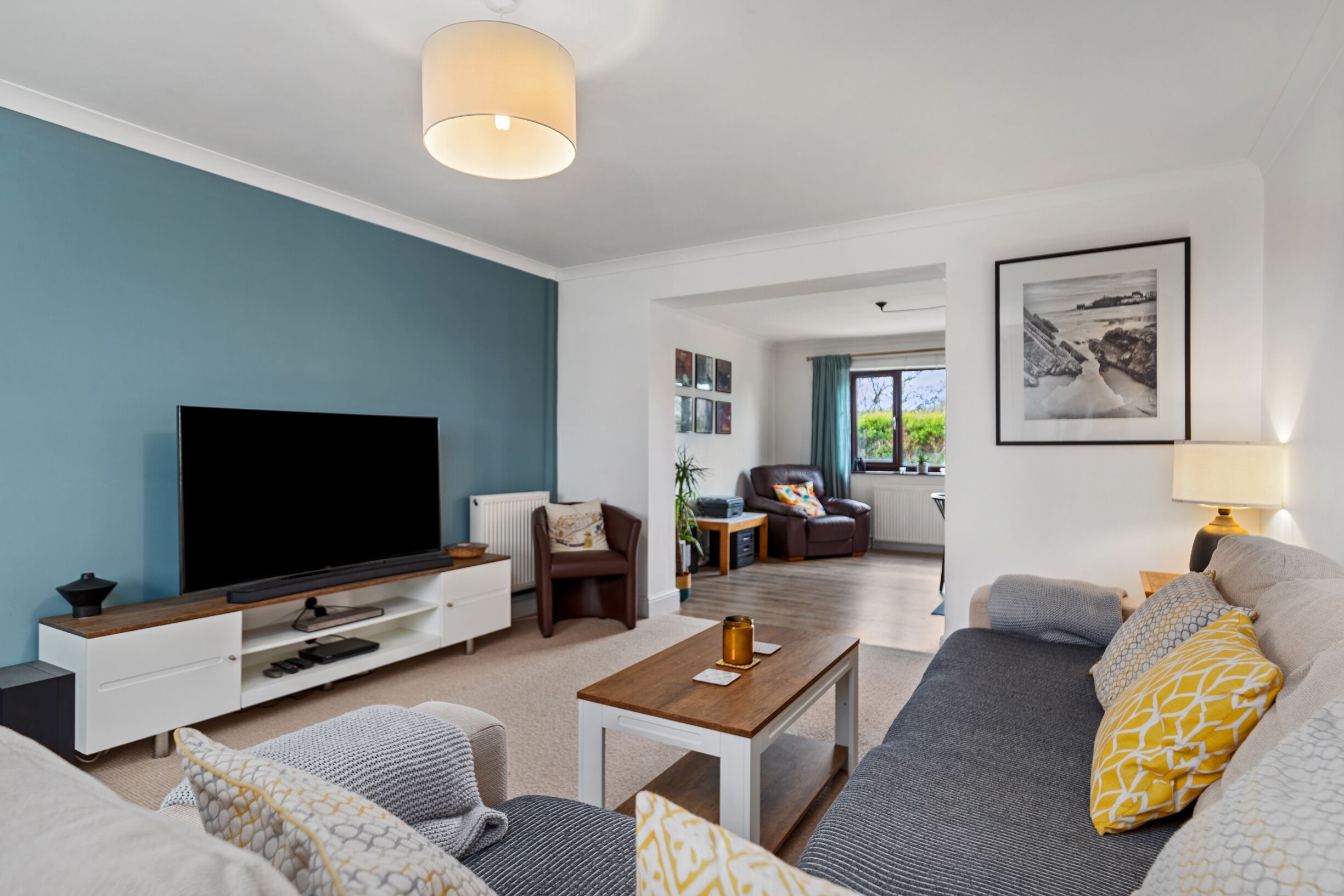 A modern living room with a flat-screen TV, a sofa, a coffee table, and a dining area with a lamp.