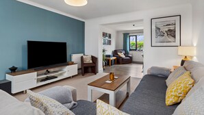 A modern living room with a flat-screen TV, a sofa, a coffee table, and a dining area with a lamp. - Woodfield House - Beautiful Tenby Cottage (Tenby)