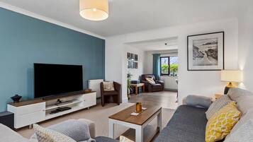 A modern living room with a flat-screen TV, a sofa, a coffee table, and a dining area with a lamp.