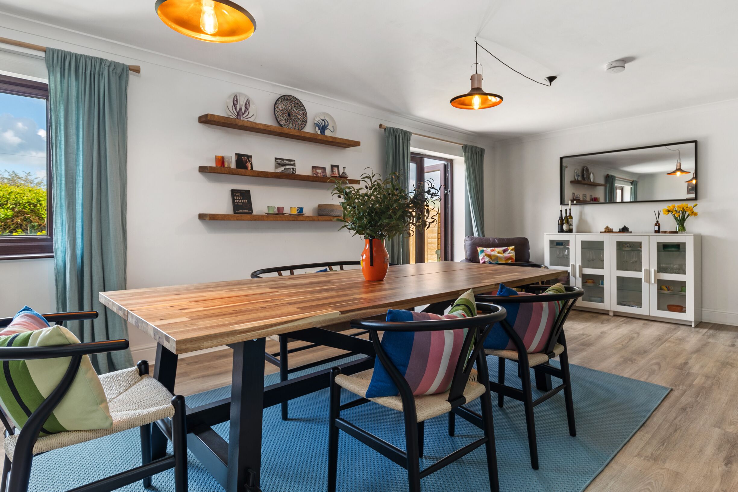 A dining area with a wooden table, chairs, and a potted plant. There are shelves with decorative items and a cabinet with glass doors.