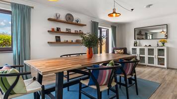 A dining area with a wooden table, chairs, and a potted plant. There are shelves with decorative items and a cabinet with glass doors.