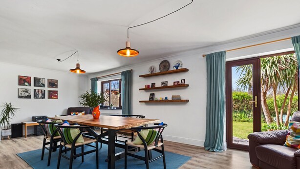 A modern dining area with a wooden table, chairs, and pendant lighting. There are shelves with decorative items and a view of the outdoors through large windows. - Woodfield House - Beautiful Tenby Cottage (Tenby)