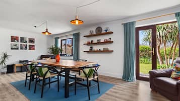 A modern dining area with a wooden table, chairs, and pendant lighting. There are shelves with decorative items and a view of the outdoors through large windows.