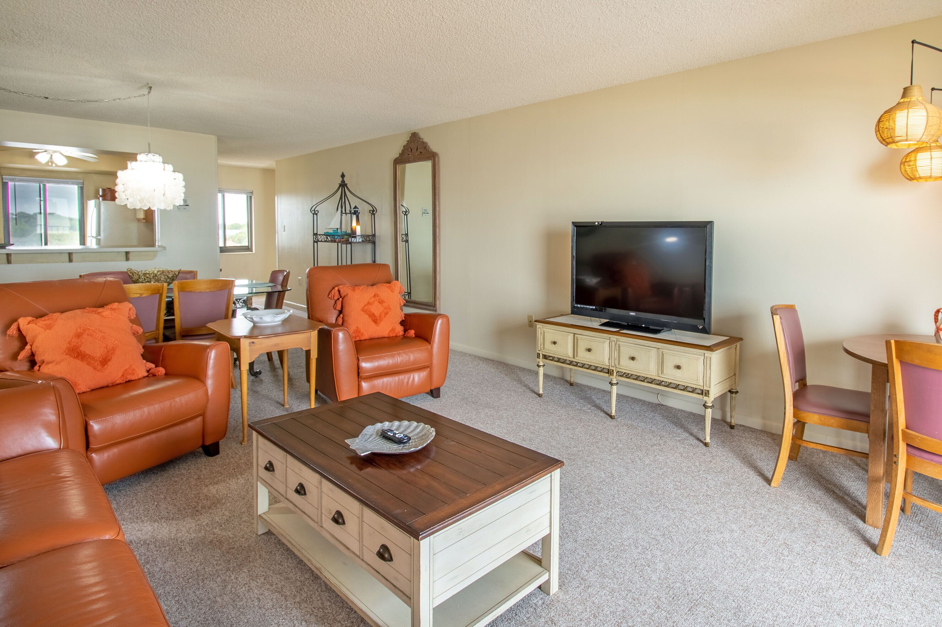 A living room with a brown leather sofa, orange cushions, a wooden coffee table, a television, and a dining area with a wooden table and chairs.