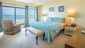 Habitación de hotel con un gran ventanal que ofrece vistas a la playa, una cama con colcha color turquesa, un banco y dos lámparas de noche.