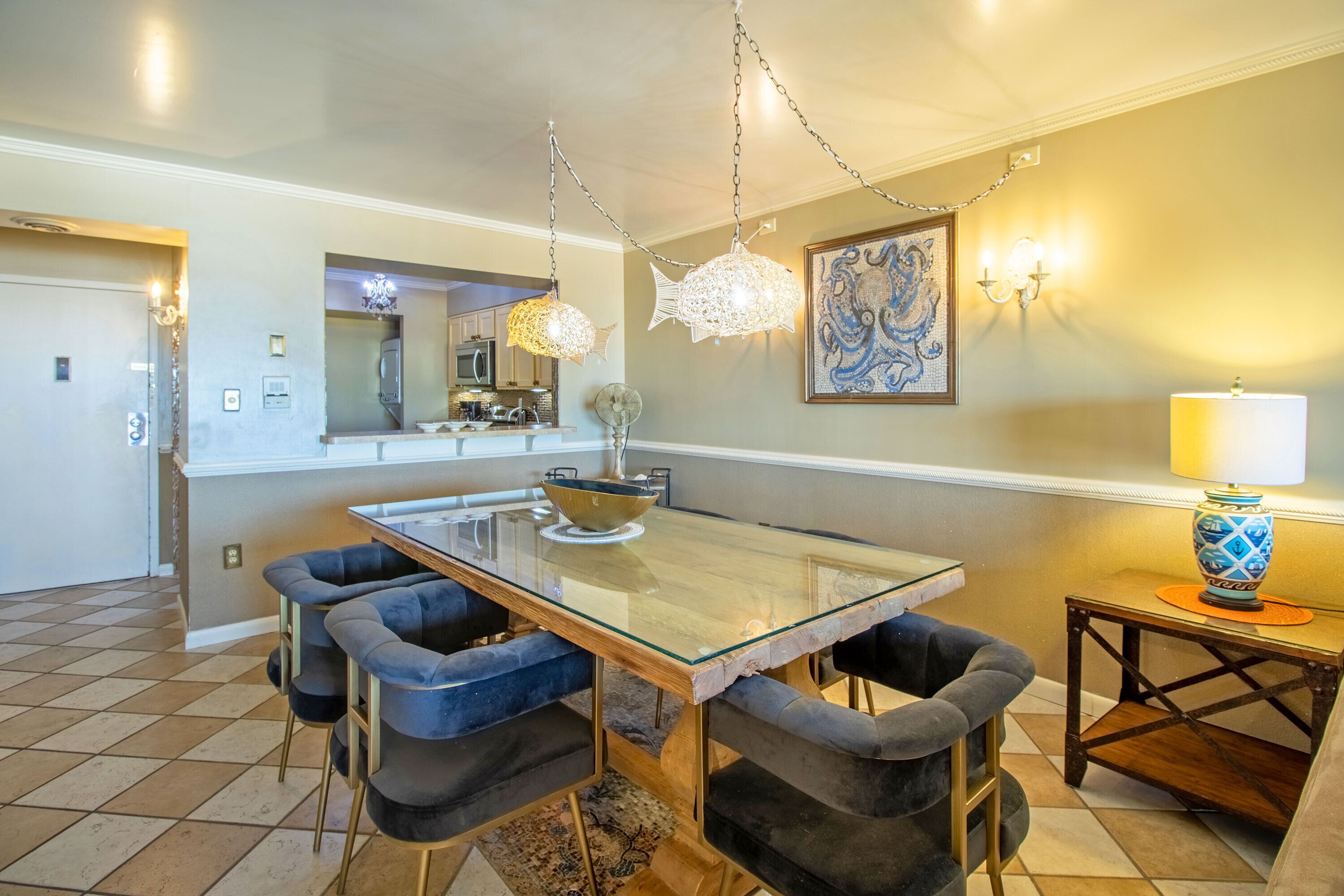 A dining area with a glass-top table, dark upholstered chairs, a chandelier, and a decorative wall piece.