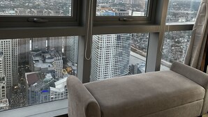Quarto em arranha-céu com grande janela e vista para a cidade, sofá bege e ventilação em metal.