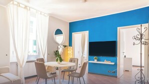 A modern living room with a round dining table, beige chairs, a sofa, a TV, and a blue accent wall.