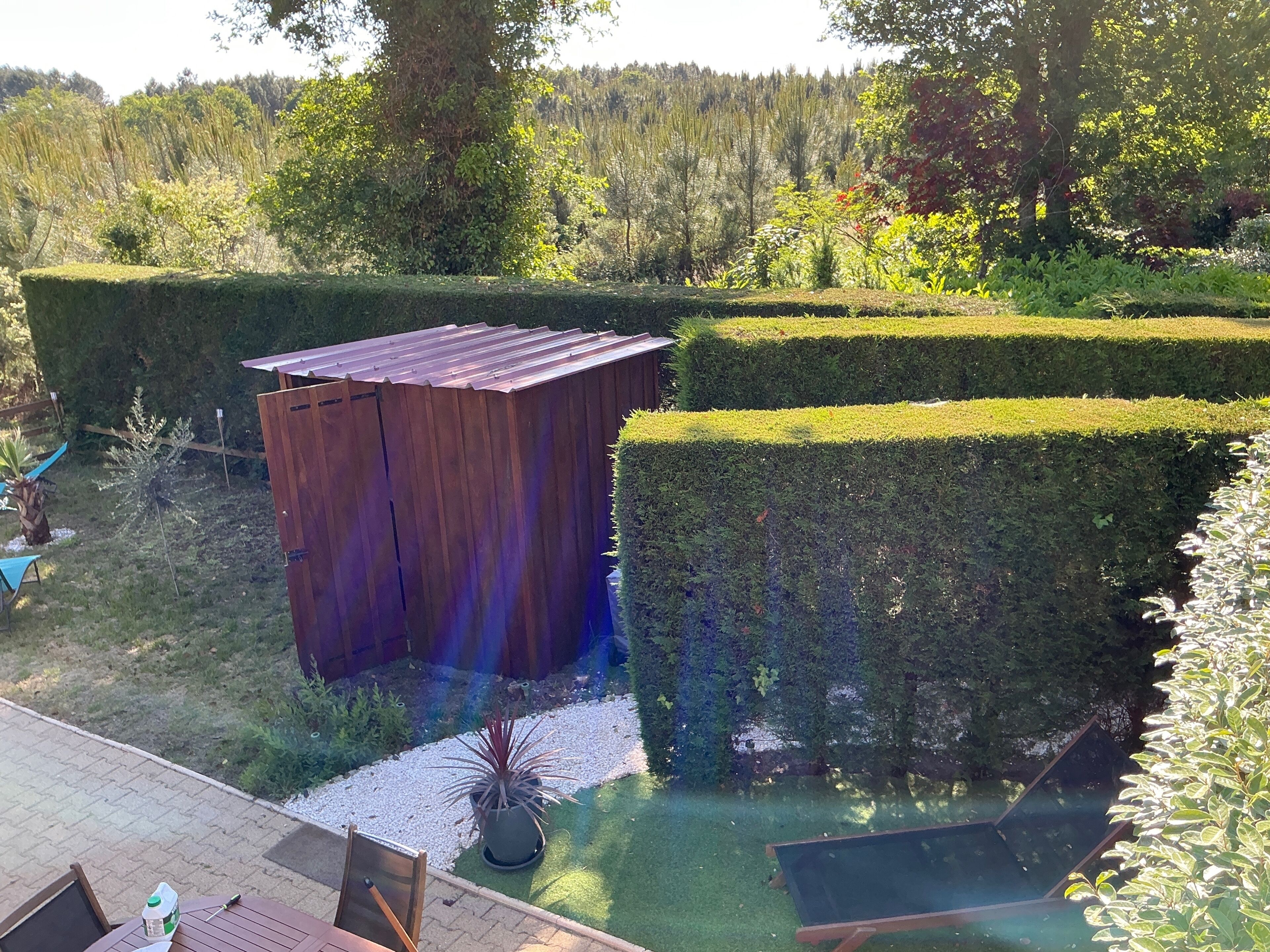 Un jardín bien cuidado con setos recortados, un cobertizo de madera y una zona de estar con mesa y sillas.