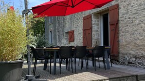 Un patio al aire libre con mesa y sillas de madera, un paraguas rojo y un edificio de piedra al fondo.