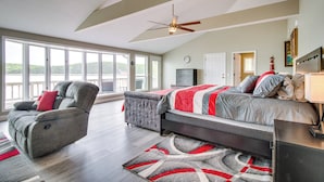 A spacious bedroom with a large bed, a recliner, and a view of the outdoors through large windows. - Serene Lakefront Mountain Home Getaway w/ Atrium! (Mountain Home)