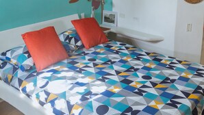 Un dormitorio con una cama, dos almohadas rojas, una colcha de estampado geométrico, un aire acondicionado de pared, un cuadro enmarcado y un adorno en la pared.