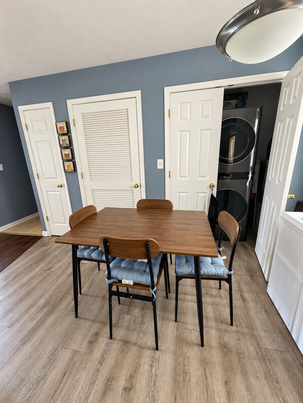 A dining area with a wooden table and four chairs, a white door, and a hallway leading to a kitchen.